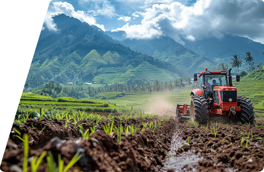 A red tractor working in a tilled field with lush green terraced mountains in the background under a cloudy blue sky.