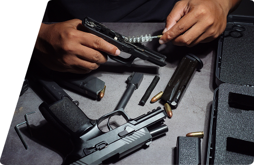 Person cleaning a disassembled handgun with a brush, surrounded by parts, magazines, and ammunition on a work surface.