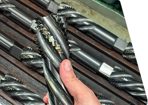 Hand holding a large, metal helical cutting tool or industrial end mill, with other similar tools resting in a rack.